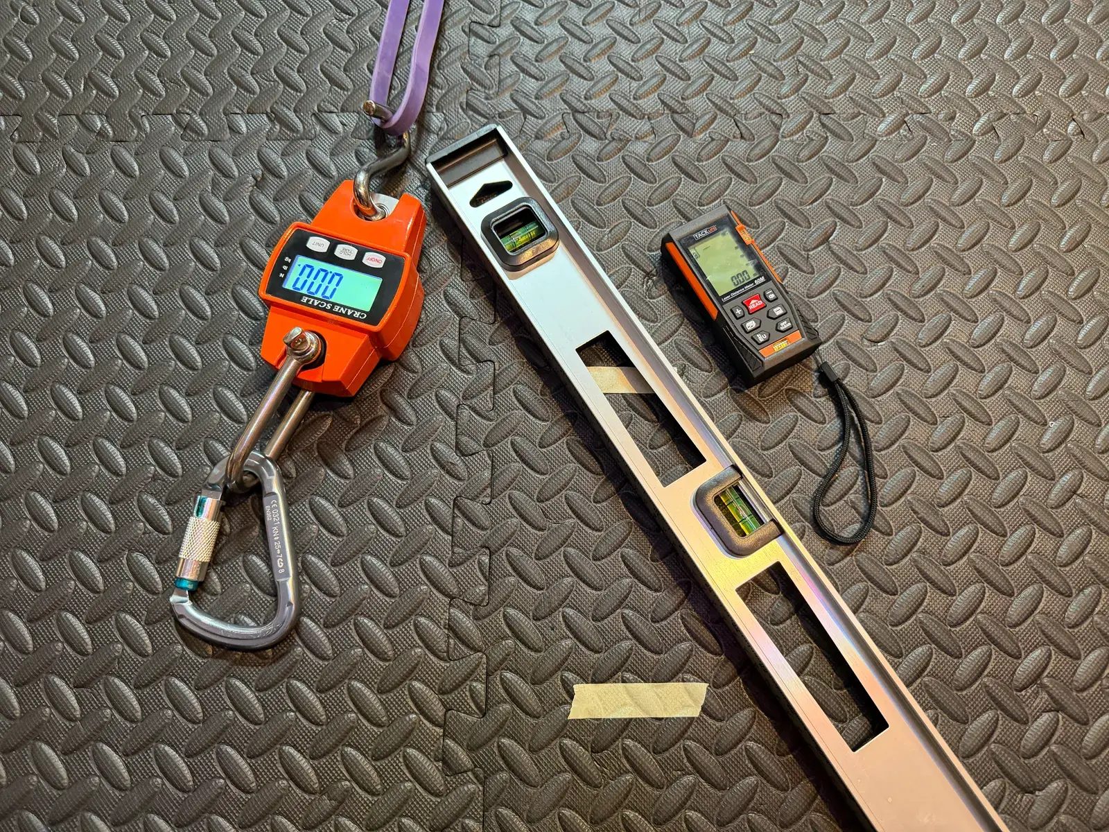 The test rig: a digital crane scale on a carabiner, a purple loop band, and a laser distance meter laid out on a gym floor